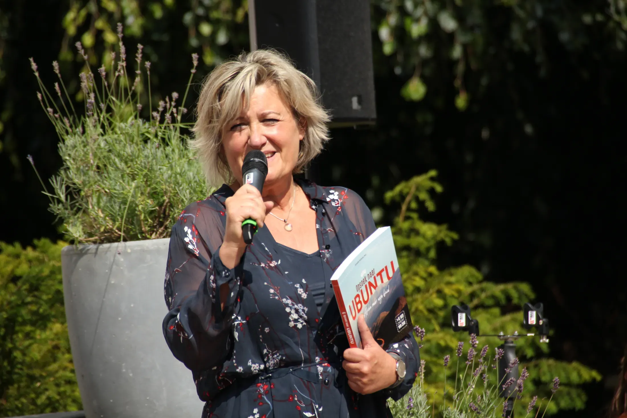 Annette Nobuntu Mul spreekt met een microfoon in de hand, haar boek Opsoek naar Ubuntu in de andere.